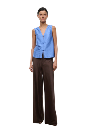 A model stands against a plain white background wearing a sleeveless light blue vest with a V-neck and button front, paired with dark brown wide-leg trousers.