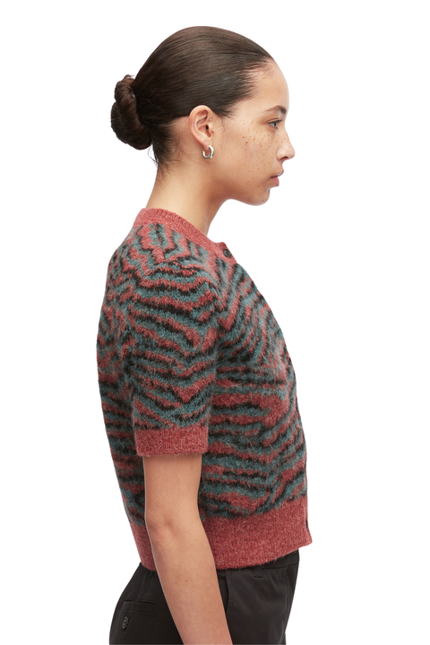 A woman shown in side profile wearing a short-sleeve fuzzy knit sweater with a wavy zebra-like pattern in muted red and dark teal, paired with black pants against a plain light gray studio background.