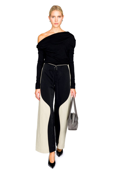 Woman posing wearing a black long sleeve off shoulder top and black and cream wide leg pants and a gray handbag on a white background