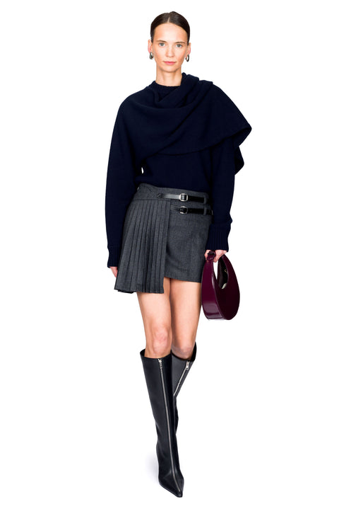 Woman posing wearing a dark blue sweater, gray pleated mini skirt, holding a red handbag and black knee-high boots on a white background