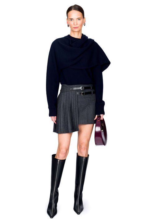 Woman wearing a navy blue draped sweater, gray pleated mini skirt, red handbag and black knee-high boots on a white background posing with one leg in front