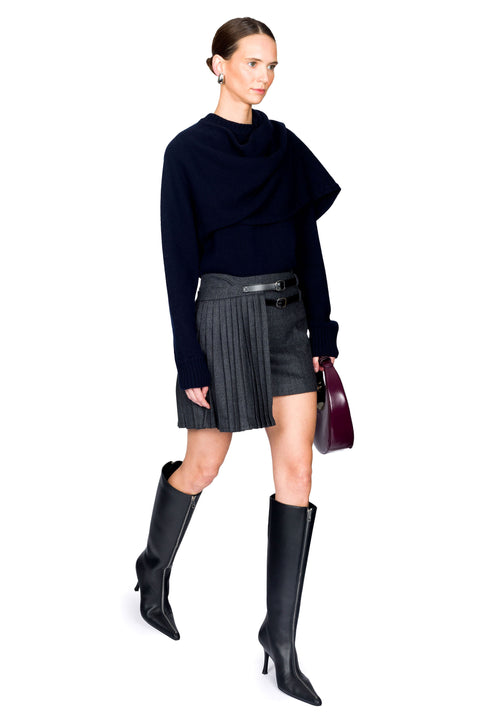 Woman walking wearing a blue sweater, asymmetrical mini pinstripe skirt, and black knee-high boots on a white background from the side