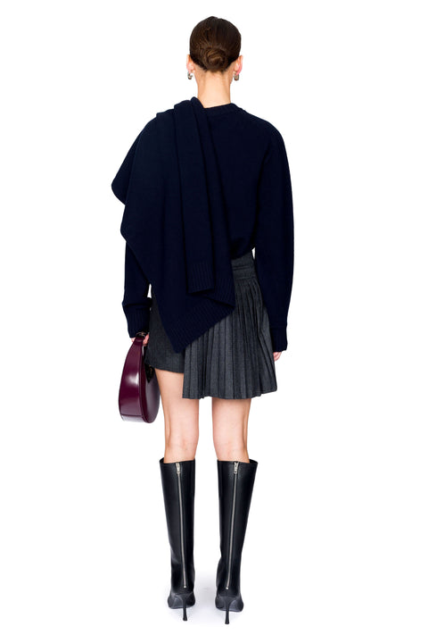 Woman facing away wearing a navy sweater, pleated skirt, and black boots on a white background