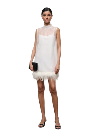 A model stands against a white background wearing a sleeveless white dress with a sheer high-neck overlay and a fluffy feather-trimmed hem. She is holding a small black clutch and wearing black strappy heels.