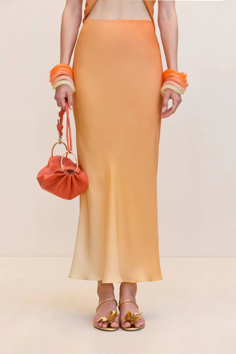 Close up of a woman wearing an orange ombre maxi slip skirt, paired with gold sandals and orange bangles. 