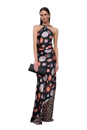 A woman modeling a sleeveless halter-neck maxi dress with a black base and a colorful circular concentric pattern in red, orange, white, and blue. She is holding a black clutch against a plain white background.