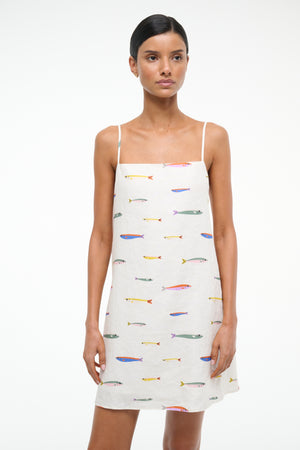 Close up of a woman wearing a spaghetti strap, linen mini dress that features a colorful fish design over a cream background. 