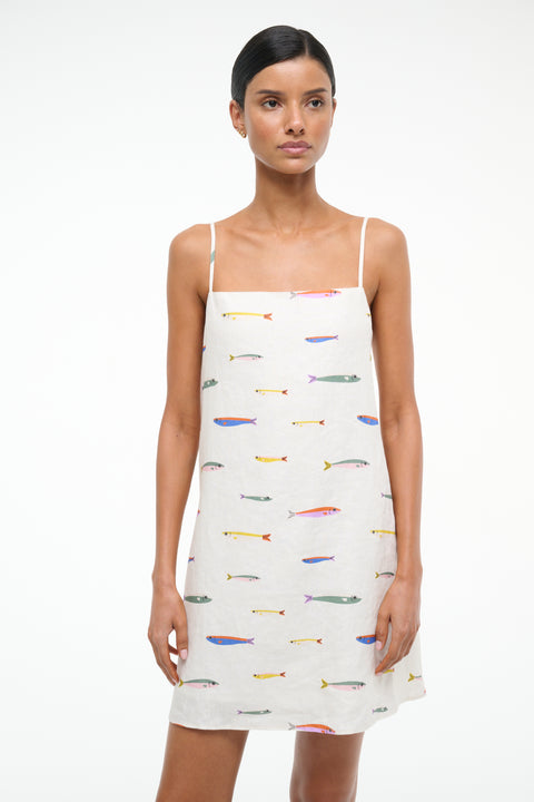 Close up of a woman wearing a spaghetti strap, linen mini dress that features a colorful fish design over a cream background. 