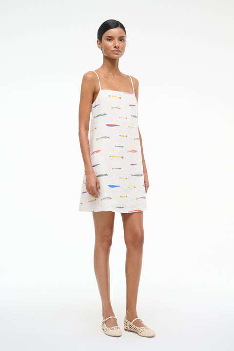 Side view of a woman wearing a spaghetti strap, linen mini dress that features a colorful fish design over a cream background. 
