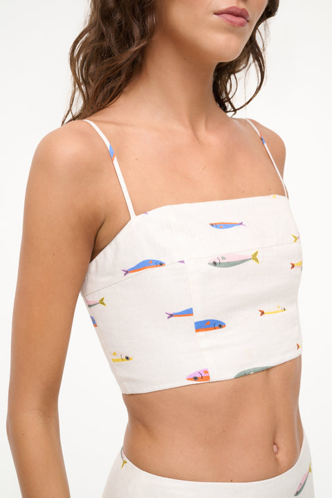 Detail shot of a woman wearing a cream spaghetti strap crop top that features a colorful fish design. 
