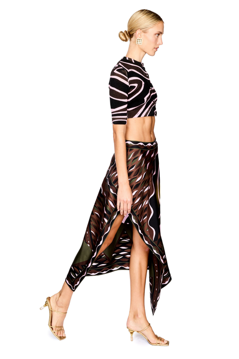 A model is shown in profile wearing a matching two-piece outfit consisting of a short-sleeve crop top sweater and a high-low skirt, both featuring a wavy abstract pattern in black, brown, and pink and green. She is wearing gold strappy high-heeled sandals against a plain white background.