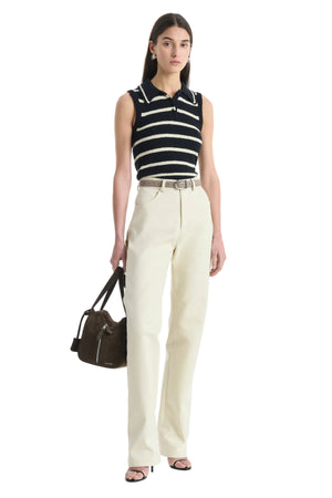 Woman wearing a black and white striped sleeveless top with cream pants, holding a brown handbag on a white background.