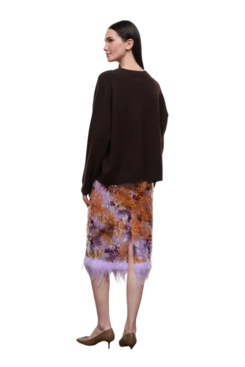 A model shown from the back wearing a dark brown knit sweater and a patterned midi skirt with purple feathers at the hem, paired with brown pointed-toe heels, against a plain white background.