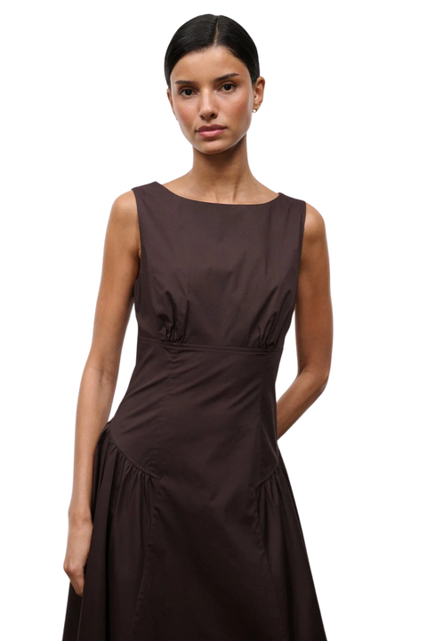 A woman modeling a sleeveless dark brown dress with a boat neckline and gathered waist details, photographed against a plain white studio background.