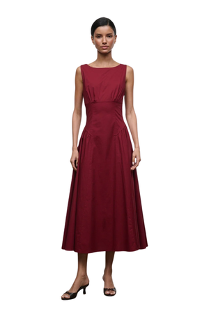 A woman modeling a sleeveless, boat-neck midi dress in deep burgundy with a structured waist and flared skirt, paired with black heeled sandals, against a plain white background.