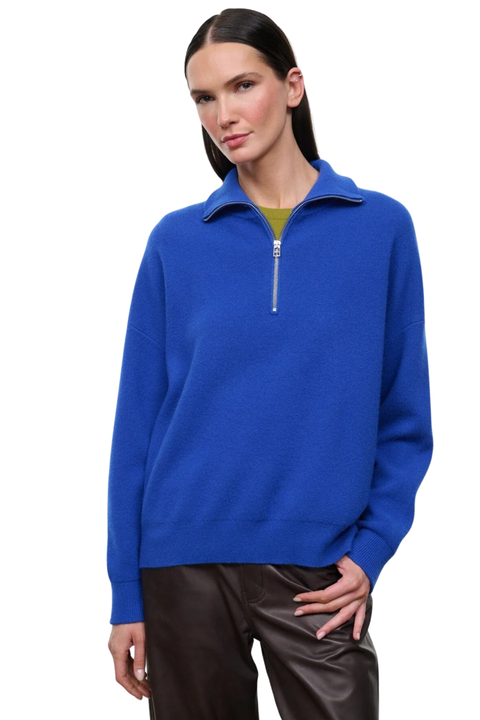 A woman modeling a bright royal blue quarter-zip pullover sweater with a stand collar, worn over a muted olive green top and paired with dark brown pants, against a plain white background.
