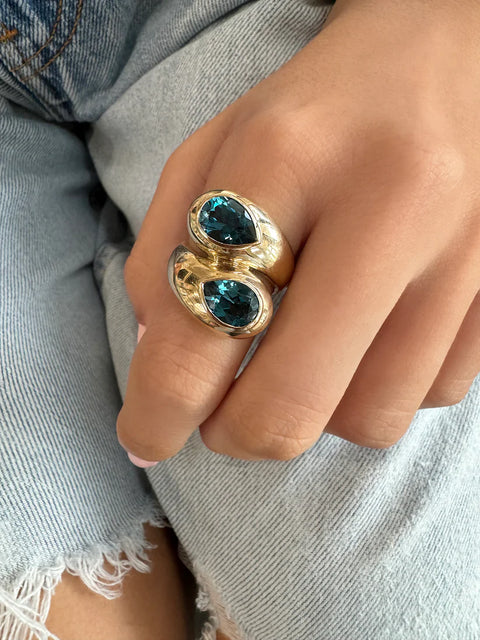 A chunky gold toi-et-moi ring that features two pear shaped blue topaz stones in a thick bezel setting, worn on a woman's hand. 