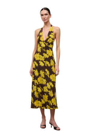 A woman modeling a sleeveless halter-neck midi dress with a bold yellow tropical floral print on a dark brown background, paired with black heeled sandals, set against a plain white studio background.