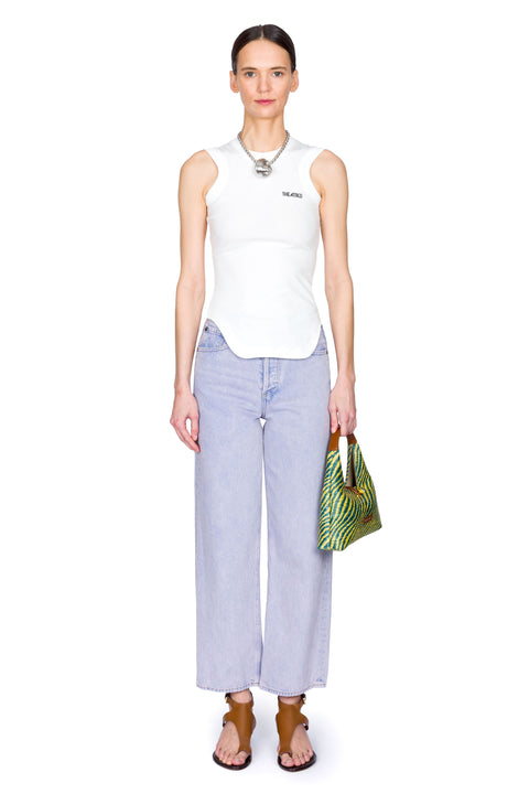 Woman wearing a white sleeveless top and high-rise, light blue jeans, holding a yellow and blue bag on a white background facing front
