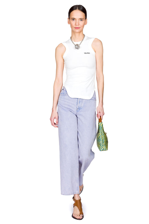 Woman wearing a white sleeveless top and high-rise, light blue jeans, holding a yellow and blue bag on a white background with one foot in front of the other