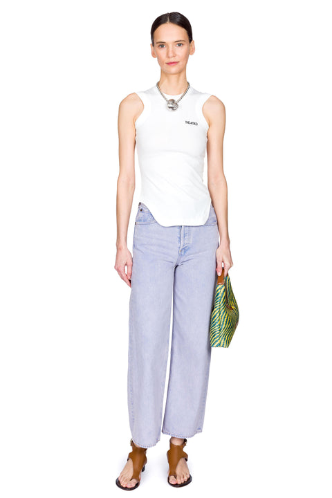 Woman wearing a white sleeveless top and high-rise, light blue jeans, holding a yellow and blue bag on a white background leaning to one side