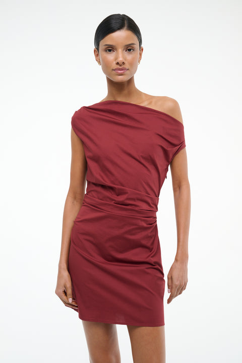 Close up of a woman wearing a deep red mini dress that features an asymmetrical off-the-shoulder neckline and draped ruching throughout, paired with black kitten heels. 