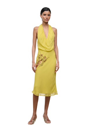 A model wearing a yellow halter-neck midi dress with a draped neckline and a lobster print on the skirt, paired with metallic wedge sandals, set against a plain white background.