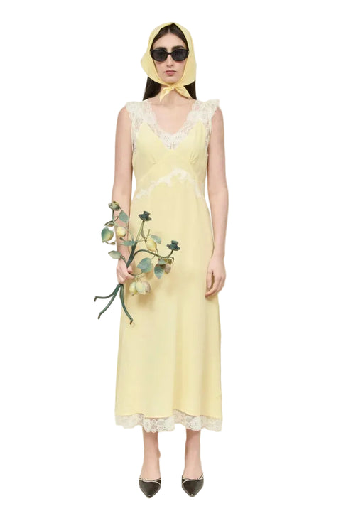 Woman wearing a yellow dress with lace details and a matching headscarf, holding flowers against a white curtain background.