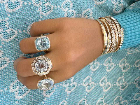 A woman wearing a large blue stone statement ring, a large aquamarine and diamond ring, and another blue stone statement ring on three fingers and five different gold and diamond bangles on her wrist. 