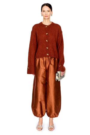 A model stands against a white studio background wearing a rust-orange cropped button-front cardigan and matching satin-like wide-leg pants in a warm copper-brown tone, with silver heeled sandals and a small patterned clutch.