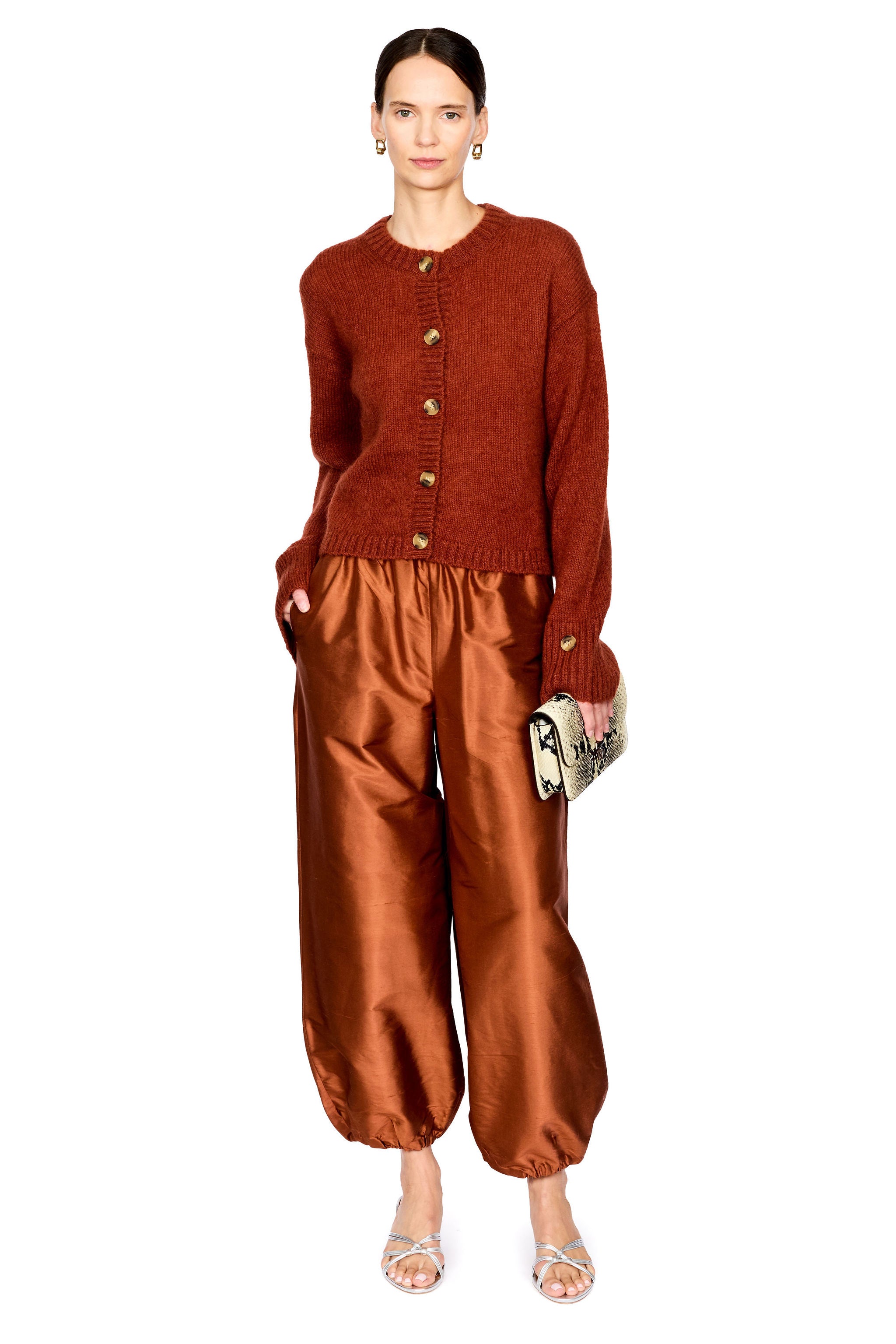 A model stands against a white studio background wearing a rust-orange cropped button-front cardigan and matching satin-like wide-leg pants in a warm copper-brown tone, with silver heeled sandals and a small patterned clutch.