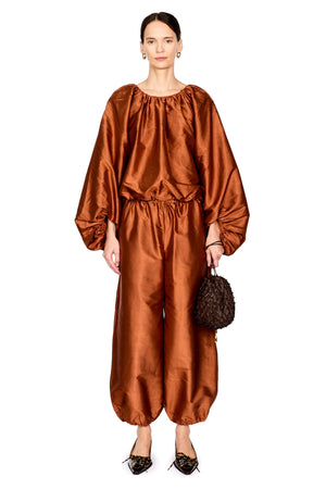 A model stands against a white background wearing a metallic copper-brown satin two-piece outfit with voluminous balloon sleeves and gathered harem-style pants, holding a small dark woven handbag.