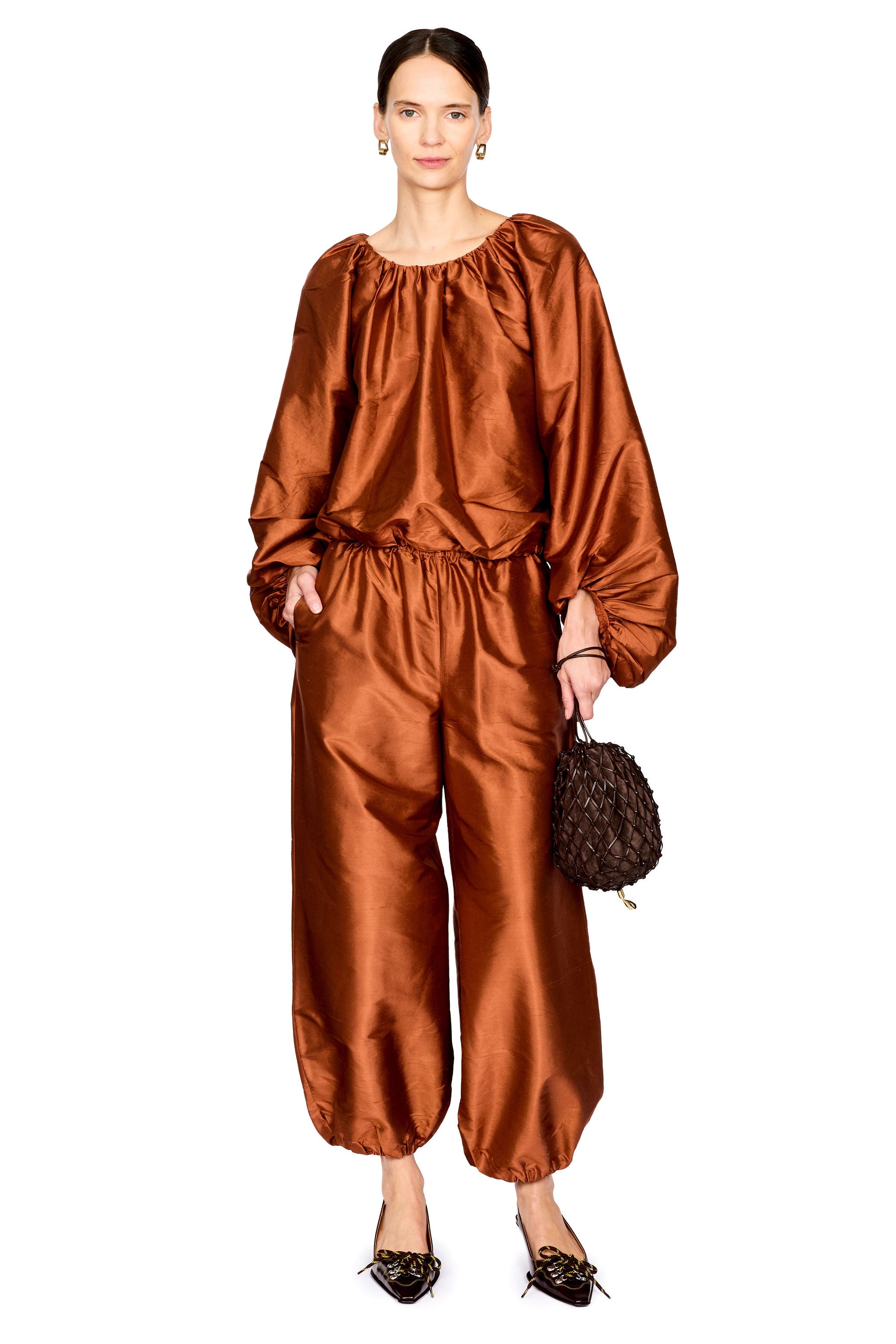 A model wearing a shiny rust-brown satin two-piece set consisting of a loose long-sleeve top and matching wide-leg pants, holding a small dark textured handbag against a plain white background.