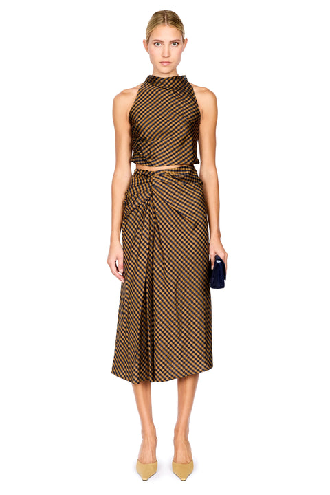A model stands against a white background wearing a brown and navy checkered halter crop top and matching draped midi skirt, paired with beige pointed-toe heels and holding a black clutch.