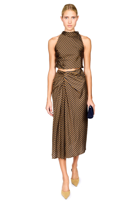 A model stands against a white background wearing a brown and navy checkered halter crop top and matching draped midi skirt, paired with beige pointed-toe heels and holding a black clutch.