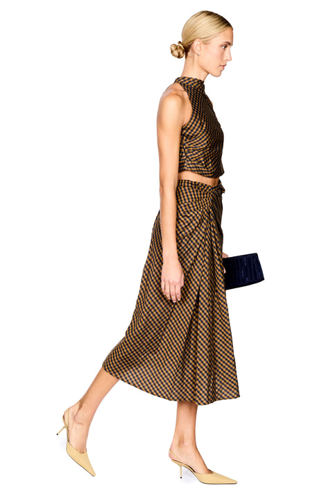 A model stands against a white background wearing a brown and navy checkered halter crop top and matching draped midi skirt, paired with beige pointed-toe heels and holding a black clutch.