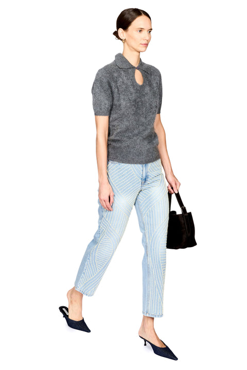 A model stands against a white background wearing a dark gray short-sleeve knit top with a keyhole cutout, light blue jeans with pearl detail, dark navy pointed shoes, and holding a dark brown handbag.