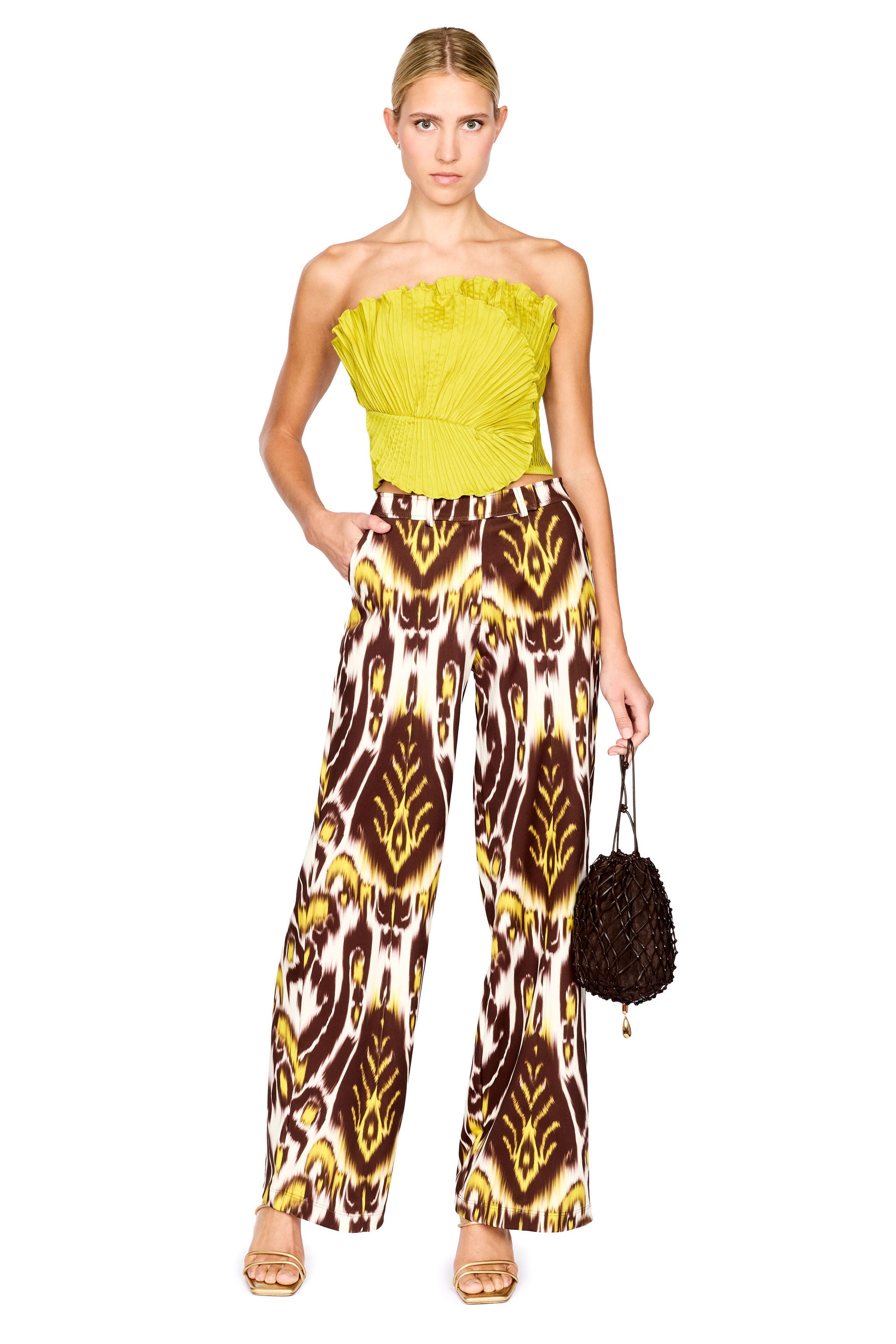 A model stands against a white background wearing a strapless bright lime top and wide-leg trousers with a brown, yellow, and white ikat pattern. She is holding a small dark brown textured handbag.