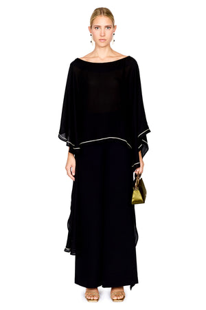 A woman modeling a black pants and a matching top with wide draped sleeves and thin white piping accents, holding a small green handbag against a plain white background.