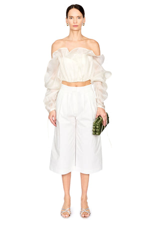 A model stands against a white studio background wearing a white off-the-shoulder puff-sleeve crop top and matching white wide-leg cropped pants, holding a small green patterned clutch.