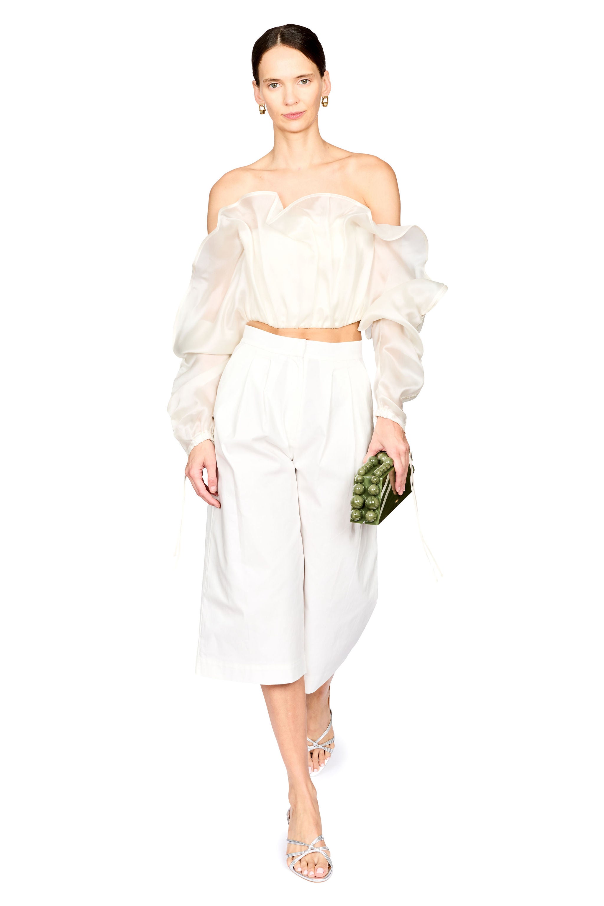 A model wearing a white two-piece outfit consisting of a sheer off-the-shoulder ruffled crop top and matching white wide-leg cropped pants, holding a green patterned clutch against a solid white background.