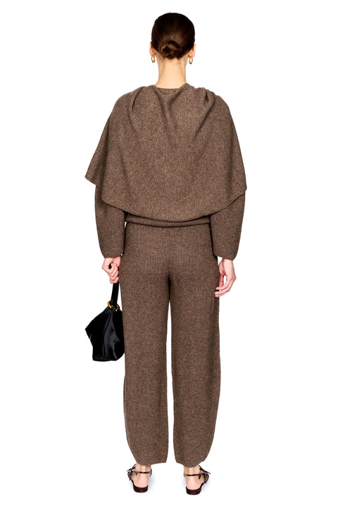 A model shown from the back wearing a matching brown knit set consisting of a draped long-sleeve top and wide-leg pants, holding a small black handbag against a plain white background.