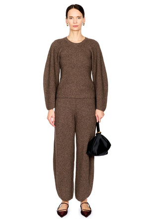 A model stands against a plain white background wearing a matching brown knit long-sleeve top and wide-leg trousers set, holding a small black handbag.