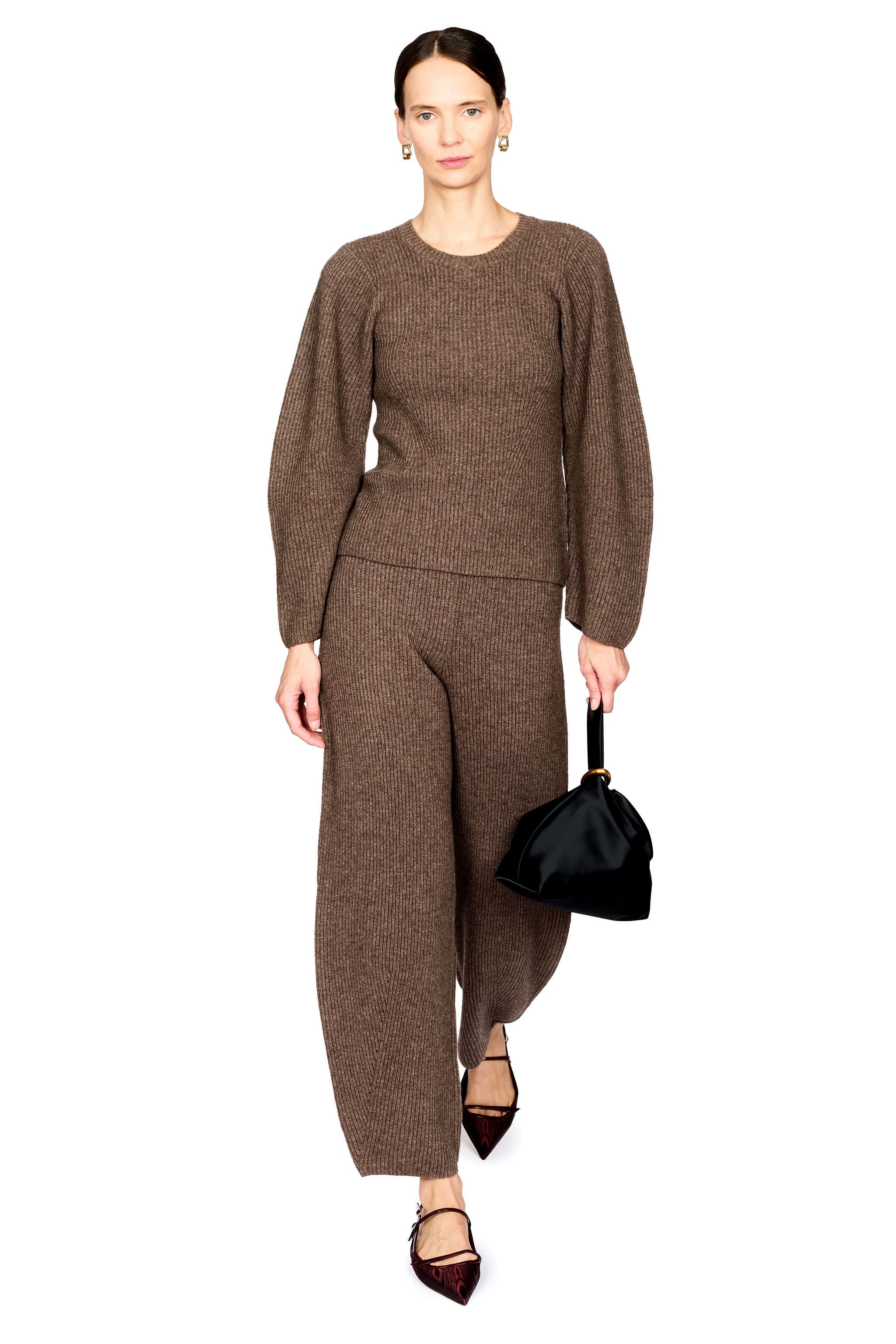A model wearing a matching brown knit two-piece set consisting of a long-sleeve crewneck sweater and wide-leg trousers, holding a small black handbag against a solid white background.