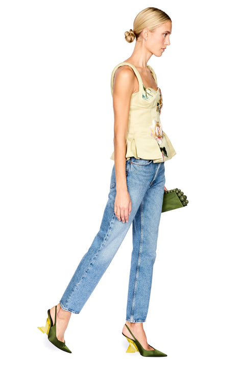 A model stands against a white background wearing a pale yellow-green floral camisole top, blue jeans, and dark green heels, while holding a small green clutch.