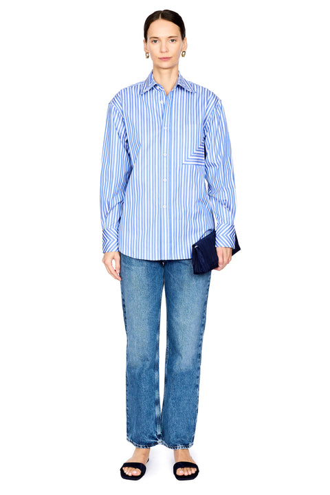 A model stands against a white background wearing a blue and white vertically striped button-down shirt, blue jeans, and black slide sandals, while holding a dark navy clutch.