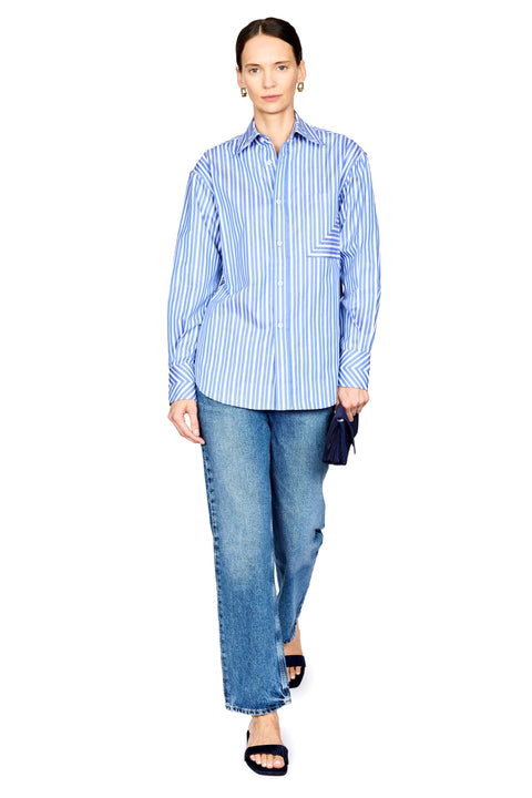 A woman modeling a blue and white vertically striped button-up shirt with blue jeans and black slide sandals, holding a small dark clutch against a plain white background.