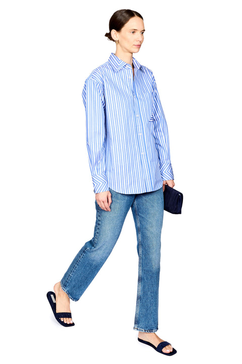 A woman modeling a light blue and white vertically striped button-down shirt, paired with blue jeans and black slide sandals, holding a small black clutch against a plain white background.