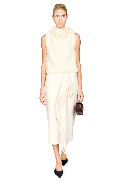 A model wearing a cream-colored sleeveless knit vest and matching midi skirt set with fringe details, paired with dark navy pointed-toe heels and a small brown handbag, against a solid white background.