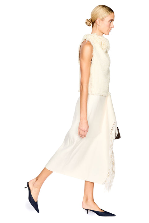 A woman modeling a sleeveless cream knit top and matching midi skirt with fringe details, paired with dark navy pointed-toe mule heels, against a solid white background.